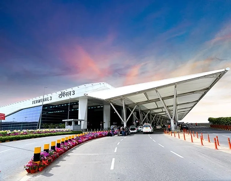 Passengers walking from IGI Airport Terminal 3 arrivals through the enclosed footbridge toward the MLCP parking and pickup zone at Pillars 4–8